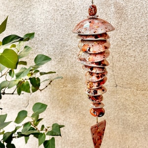 May include: A hanging ceramic mobile featuring a series of stacked, rounded, and speckled pink and orange discs suspended on a cord. The mobile terminates in a single triangular ceramic piece at the bottom, all finished with a glossy glaze.