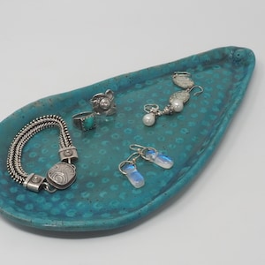 May include: A collection of jewelry displayed on a turquoise ceramic tray. The jewelry includes a silver bracelet, rings with turquoise and clear stones, pearl earrings, and iridescent earrings. The tray has a dotted pattern.