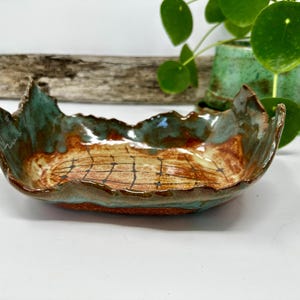 May include: A handmade ceramic bowl with a unique, organic shape. The bowl features a blend of colors, including teal, brown, and orange, with a grid-like pattern in the center. The rim has a jagged edge, adding to its artistic appeal.