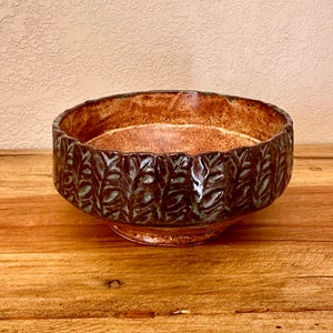 May include: A handcrafted ceramic bowl with a textured leaf pattern around the rim. The bowl features a brown interior and a dark brown exterior with green accents. The bowl sits on a small, round base and is displayed on a wooden surface.