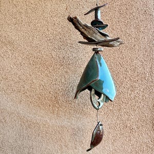 May include: A handcrafted wind chime with a teal ceramic bell, driftwood, and metal accents. The bell has a glossy finish and hangs below a cluster of wooden and metal elements. The overall design is organic and artistic.