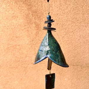 May include: A handcrafted ceramic wind chime with a bell-shaped body in shades of teal and blue. The chime features a stack of dark blue ceramic discs and a rectangular bottom piece. The wind chime hangs from a thin, natural-colored cord.