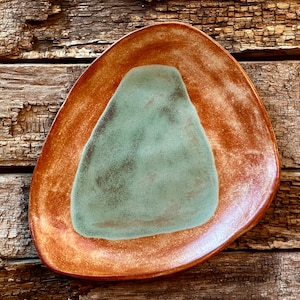 May include: A ceramic plate with a unique, irregular shape. The plate is glazed in a light brown color with a large, irregular, teal-colored area in the center.