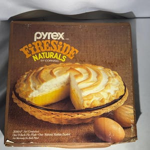May include: A Pyrex Fireside Naturals by Corning product box featuring a clear glass pie plate inside a natural rattan basket. The box displays a baked pie with a golden yellow filling and a browned meringue topping, along with two brown eggs.