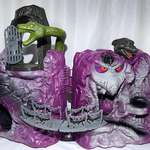 May include: A purple and grey plastic toy playset featuring a large sculpted face with orange eyes, a green snake figure extending from a dark archway, a grey gate, and a bridge connecting two sections of the textured, rocky-looking structure.