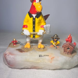 May include: A hand-painted figurine featuring a red rooster dressed as a firefighter in a yellow jacket and black helmet, holding a silver axe. Beside it is a smaller bird figure, a red fire hydrant, and a small wooden house with orange flames.