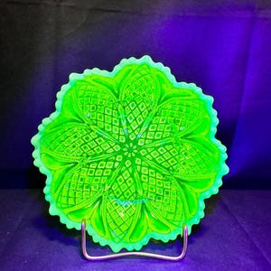 Puede incluir: Un plato de vidrio prensado de color verde neón brillante y luminoso, con un patrón geométrico complejo y simétrico de diamantes y pétalos radiantes. El plato circular tiene un borde festoneado y ondulado sobre un soporte.