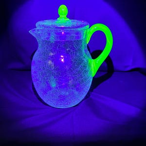 Op de afbeelding: Een glazen kan met een craquelé textuur en een deksel, die onder ultraviolet licht fel neongroen oplicht. De kan heeft een bolvormig lichaam, een kleine tuit en een gebogen handvat, waarbij het handvat en de dekselknop intens oplichten.