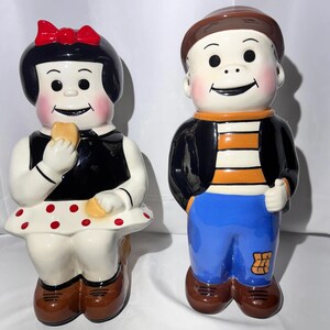 May include: Two ceramic figurines depicting a pair of children. The left figure has black hair with a red bow, a black top, and a white skirt with red polka dots. The right figure wears a brown cap, a black and orange striped shirt, and blue pants.