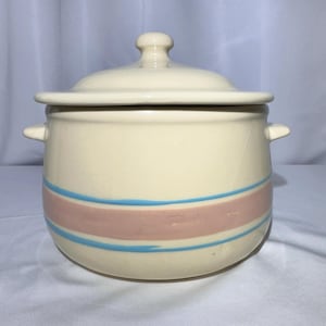 May include: A cream-colored ceramic pot with a lid and two side handles. The pot features a wide horizontal band in a muted pink hue, bordered by two thin, hand-painted blue stripes that wrap around the center of the rounded vessel body.