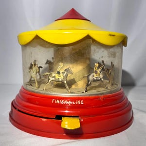May include: A vintage toy carousel featuring a bright red base with the text FINISH LINE printed in white. The toy has a yellow scalloped roof with a red pointed top. Inside a clear plastic enclosure, several small horse figures with riders.