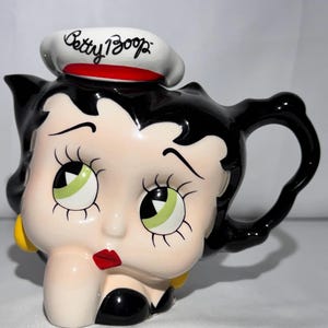 Puede incluir: Una tetera de cerámica con forma de cabeza de Betty Boop, con cabello negro, ojos verdes, labios rojos y pendientes amarillos. La tapa es un gorro de marinero blanco con una banda roja y el texto Betty Boop escrito en negro en el frente.