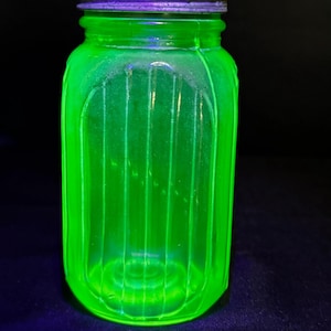 Puede incluir: Un frasco de vidrio de color verde neón brillante con detalles acanalados verticales en los lados. El recipiente está rematado con una tapa metálica circular y texturizada que se ve de color morado oscuro bajo la luz ultravioleta.