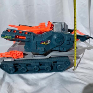 May include: A dark blue and gray plastic toy tank with bright orange missile launchers and cannons. The side features a sticker with the text BRAWLER and a missile lock danger blast warning. A yellow measuring tape shows a height of 8 inches.