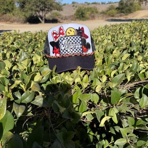 Puede incluir: Una gorra tipo camionero en blanco y negro con un parche central de patrón de cuadros blanco y negro y la palabra Mama en letra cursiva. La gorra está decorada con varios parches que incluyen un lazo rojo, una cara sonriente y estrellas rojas.