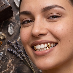 May include: A set of four gold tooth caps, known as grills, worn on the upper and lower teeth. The gold caps are smooth and polished, covering individual teeth to create a metallic appearance. The teeth are visible in a smiling mouth.