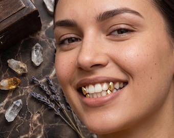 May include: A set of four gold tooth caps, known as grills, worn on the upper and lower teeth. The gold caps are smooth and polished, covering individual teeth to create a metallic appearance. The teeth are visible in a smiling mouth.