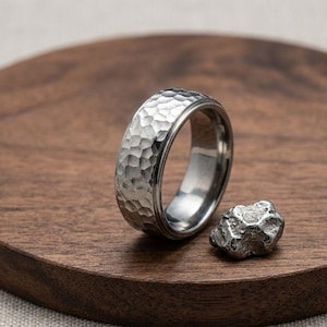 May include: A silver-toned metal ring featuring a textured, hammered exterior surface with a polished inner band, accompanied by a small, irregular, rough-hewn piece of matching metallic material positioned next to the ring.