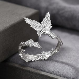 May include: A silver-colored open ring featuring a detailed phoenix design with spread wings and textured feathers. The band is adjustable and includes the engraving S925 on the inner surface, indicating sterling silver composition.