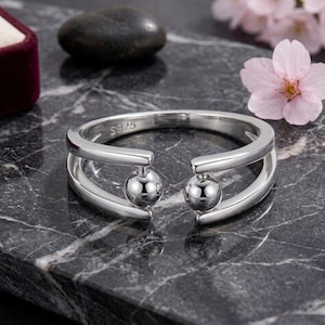 Può includere: Un anello aperto in argento con due perline sferiche lucide alle estremità della fascia. La superficie interna della fascia presenta il testo S925 chiaramente impresso, che indica la composizione del metallo. L'anello ha una finitura liscia e riflettente.