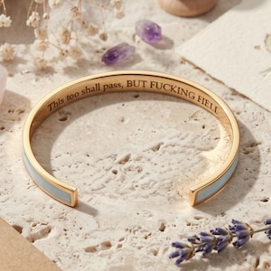 Op de afbeelding: Een goudkleurige armband met een lichtblauwe emaille inleg. De binnenzijde van de armband is gegraveerd met de zwarte tekst "This too shall pass, BUT FUCKING FIELL". De armband heeft een open ontwerp en een gepolijste metalen afwerking.