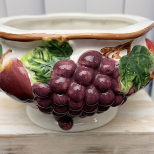 May include: A ceramic bowl featuring a raised, three-dimensional design of purple grapes and green leaves around the exterior. The bowl has a white base colour, a scalloped rim with a brown painted edge, and a pedestal foot for stability.