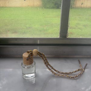 May include: A small, clear glass bottle filled with a transparent liquid, topped with a cylindrical wooden cap. A braided, tan-coloured cord with metallic gold threading is looped through the cap, featuring a single wooden bead for adjustment.