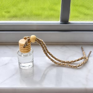 Könnte beinhalten: Eine kleine, klare Glasflasche mit Flüssigkeit, gekrönt von einem runden, hellen Holzdeckel. Eine geflochtene beige Kordel mit metallisch goldenen Fäden ist am Deckel befestigt und verfügt über eine verschiebbare Holzperle zur Anpassung sowie Metallenden.