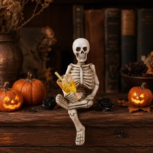 May include: A white skeleton figurine sitting with legs crossed, holding a clear glass filled with a yellow liquid and a yellow-and-white striped straw. The skeleton is positioned between two small orange jack-o'-lanterns with glowing faces.