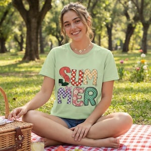 May include: A light green short-sleeved t-shirt featuring the word SUMMER printed in large, multi-patterned block letters. Each letter displays a different design, including watermelon, gingham, polka dot, and floral prints in red, orange, and green.