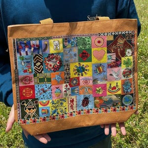May include: A tan tote bag featuring a large rectangular panel covered in a colourful patchwork of embroidered squares. The diverse designs include a ladybug, a turtle, a heart, a dove, a four-leaf clover, a rainbow, and various cultural symbols.
