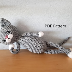 May include: A gray crocheted cat laying on a wooden surface. The cat has white paws and a pink nose. The text "PDF Pattern" is visible in the upper right corner of the image.