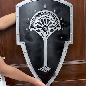 May include: A black shield with a silver metallic border featuring a series of rivets along the edge. The centre displays a silver tree-like emblem with circular patterns at the top, surrounded by seven small four-pointed star shapes.