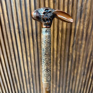Può includere: Un bastone da passeggio con un manico metallico color rame a forma di testa di coniglio con tratti del viso dettagliati e lunghe orecchie. La parte superiore dell'asta è decorata con un intricato motivo floreale inciso su legno chiaro.