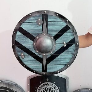 Peut inclure: Un bouclier rond en bois avec un motif de grain de bois bleu sarcelle et noir, doté d'un umbo métallique central. Le bouclier est bordé d'un rebord en métal sombre fixé par des rivets et présente un motif en losange et un croissant de lune.