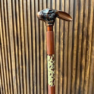 Può includere: Un bastone da passeggio con un'impugnatura metallica color bronzo a forma di testa di coniglio. L'asta è marrone con un inserto decorativo in metallo dorato che mostra un motivo a tralci floreali, accentuato da una sottile fascia dorata.