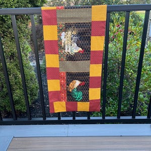 May include: A rectangular fabric table runner featuring a central black panel with a honeycomb pattern and two rooster illustrations. The border consists of alternating mustard yellow and deep red squares with a subtle eyelet embroidery texture.
