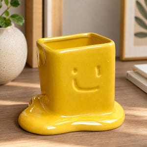 Pode incluir: Um recipiente de cerâmica amarelo brilhante com formato quadrado e acabamento lustroso. A parte frontal apresenta um design simples de rosto sorridente em relevo. A base do recipiente é esculpida para parecer uma poça de líquido.