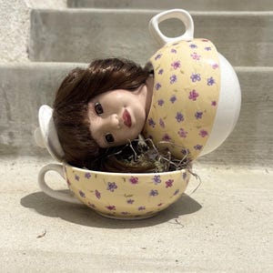 Könnte beinhalten: Ein Keramik-Puppenkopf mit braunem Haar in einer gelben Teetasse, die mit kleinen violetten und blauen Blumen verziert ist. Der Puppenkopf liegt schräg und ist teilweise von einem passenden gelben Tassendeckel mit weißem Griff bedeckt.