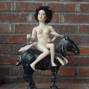 May include: A porcelain doll with short black hair and a pale complexion sits atop a black painted wooden horse figurine. The doll is adorned with light-colored feathers around the waist and shoulders, while the horse features a distressed finish.