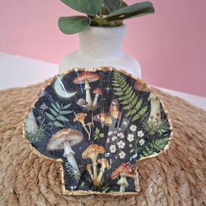 Peut inclure: Un plat décoratif en forme de coquille Saint-Jacques avec un fond sombre orné d'un imprimé botanique de champignons, fougères, petites fleurs blanches, un papillon et un croissant de lune. Les bords sont finis par un liseré doré métallique.