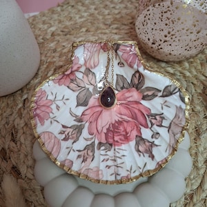 Puede incluir: Un colgante de piedra preciosa morada en forma de lágrima sobre una fina cadena dorada, que descansa dentro de una concha de vieira blanca decorada con un patrón floral rosa y bordes dorados. La concha se asienta sobre una base gris clara.