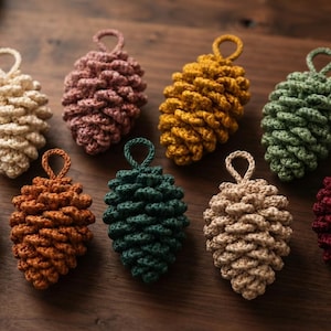 Può includere: Una collezione di nove ornamenti a forma di pigna realizzati all'uncinetto in vari colori, tra cui crema, rosa antico, giallo senape, verde salvia, lavanda, arancione bruciato, verde bosco scuro, beige e bordeaux intenso, con un occhiello.