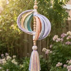 Peut inclure: Un mobile suspendu composé d'une série de bandes de tissu crochetées et incurvées dans des tons pastel de lavande, pêche, vert menthe et bleu clair. Les bandes sont fixées à une cordelette verticale de perles en bois naturel avec un long pompon.