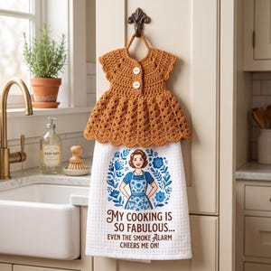 Puede incluir: Un paño de cocina blanco con textura de gofre y un adorno de vestido de ganchillo marrón con dos botones. El paño muestra un gráfico floral azul de una persona con delantal y el texto: MY COOKING IS SO FABULOUS... EVEN THE SMOKE ALARM CHEERS ME ON!