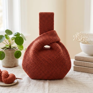 Op de afbeelding: Een roestkleurige, handgebreide tas met een textuur van rijststeek. De tas heeft een uniek knoop-handvatontwerp waarbij de ene kant langer is dan de andere, wat een asymmetrische opening creëert om de tas gemakkelijk met de hand te dragen.
