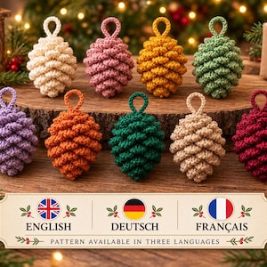 Puede incluir: Una colección de nueve adornos de piñas de ganchillo en varios colores, incluyendo crema, rosa, amarillo, verde, morado, naranja, verde oscuro, beige y rojo. Un letrero rectangular debajo muestra el texto: ENGLISH, DEUTSCH, FRANÇAIS, PATTERN AVAILABLE IN THREE LANGUAGES.