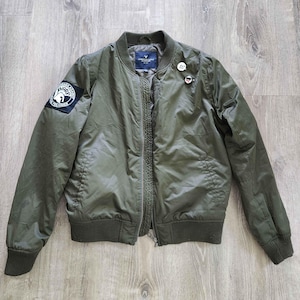 May include: An olive green bomber jacket featuring a ribbed collar, cuffs, and hem. The jacket has a front zipper closure, two side pockets, and a circular patch on the right sleeve with the text "SHARP" and "PRE-FORCE". Two pins are attached.