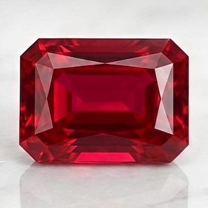 May include: A vibrant, deep red gemstone cut in a rectangular emerald shape with multiple facets that reflect light. The stone displays a rich, saturated crimson hue and a clear, transparent quality, showcasing precise geometric edges and symmetry.