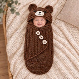 Könnte beinhalten: Eine dunkelbraune, gehäkelte Baby-Wickeldecke mit integrierter Kapuze, Bärenohren und einem Bärengesicht. Die Wickeldecke hat eine gerippte Textur und wird mit drei großen, runden, cremefarbenen Knöpfen an der diagonalen Vorderklappe verschlossen.